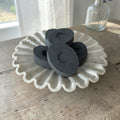 Four dark charcoal-colored oval soak stone bath bombs with crescent moon cutouts, arranged on a white scalloped ceramic plate on a rustic wooden table.